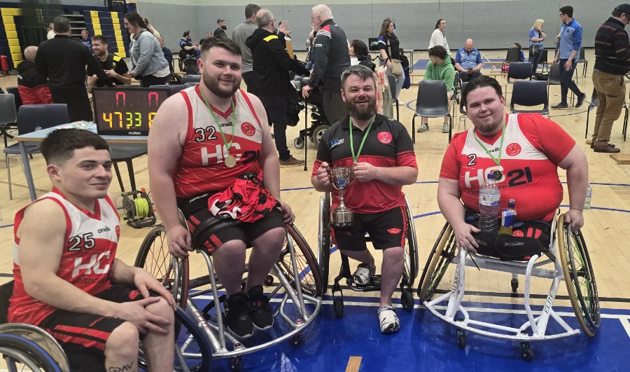 four wheelchair basketball players posing with medals and trophy on a basketball court